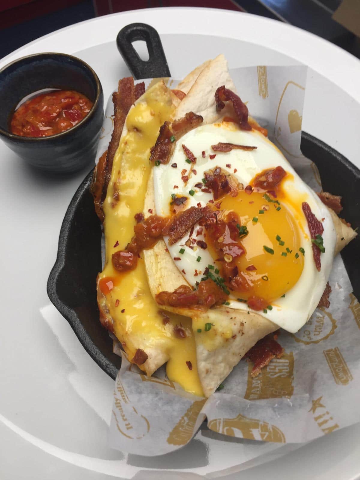 Folded tortilla topped with a fried egg, crispy bacon, cheese sauce and chilli flakes in a small cast iron pan, with a pot of harissa